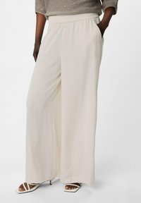 Pantaloni a gamba larga di colore crema realizzati in tessuto leggero. Presentano un cinturino elastico e tasche laterali. Abbinati a tacchi bianchi con cinturini.