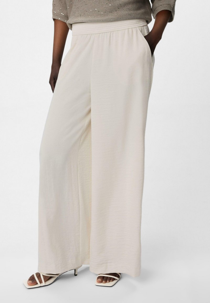 Pantaloni a gamba larga di colore crema realizzati in tessuto leggero. Presentano un cinturino elastico e tasche laterali. Abbinati a tacchi bianchi con cinturini.