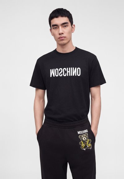 Νέος άνδρας με μαύρο t-shirt Moschino με αντίστροφο λογότυπο και μαύρο παντελόνι με αρκουδάκι και το κείμενο Moschino, στέκεται με τα χέρια στις τσέπες.