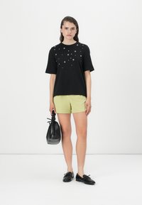 Zwarte t-shirt met korte mouwen en ronde zilveren versieringen, gecombineerd met lichtgroene shorts en een zwarte handtas met textuur.