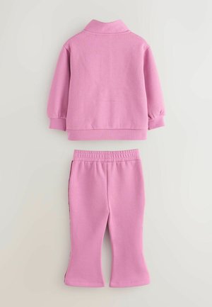 Roza dvodelna obleka, ki vključuje pulover s ovratnikom in manšetami, v kombinaciji z hlačami z elastičnim pasom in razširjenimi robovi.