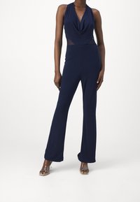 Marinblå jumpsuit med draperad halsringning, figurnära midja och utsvängda ben. Tillverkad av mjukt tyg med genomskinliga sidopaneler och minimalistisk hårdvara.