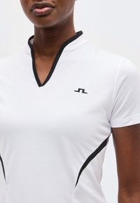 Κοντινό πλάνο ενός ατόμου που φοράει λευκό πουκάμισο με μαύρο περίγραμμα στο λαιμό σε σχήμα V και στις πλευρές, με μικρό μαύρο λογότυπο στο στήθος.