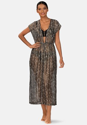 Kobieta w przezroczystej, czarno-brązowej sukience maxi z wzorem, nosząca czarne bikini, stojąca na boso z jedną ręką na biodrze.