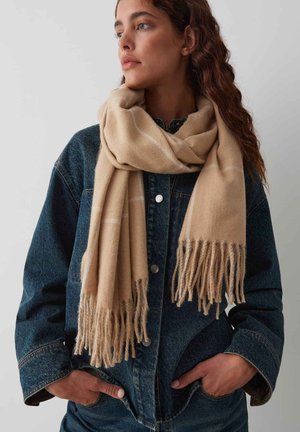 Beige scarf with fringed edges, made from soft material, wrapped around the neck, paired with a dark denim jacket.