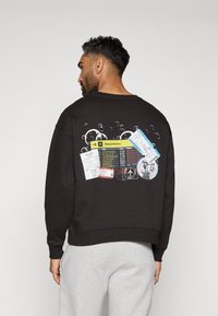 Melnā krekla ar grafiku, kas attēlo ceļojumu biļetes un izlidošanas informācijas paneli uz muguras. Izgatavots no mīkstas auduma, ar ribotām aprocēm.