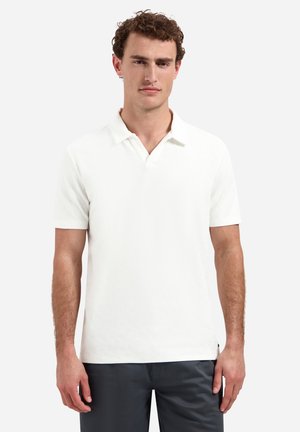 Man met krullend haar die een effen wit polo met korte mouwen en een donkere broek draagt, staand tegen een effen lichte achtergrond.