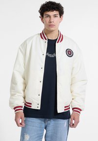 Junger Mann trägt eine weiße Collegejacke mit rot gestreiftem Kragen und Bündchen, ein schwarzes Grafik-T-Shirt und hellblaue, zerrissene Jeans.