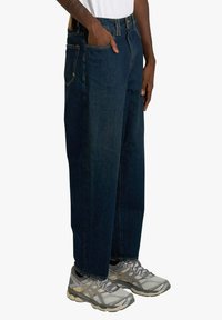 Jeans in denim blu scuro con una vestibilità comoda, dotati di tasche anteriori, chiusura con bottoni e cuciture sottili dorate, abbinati a scarpe da ginnastica grigie.