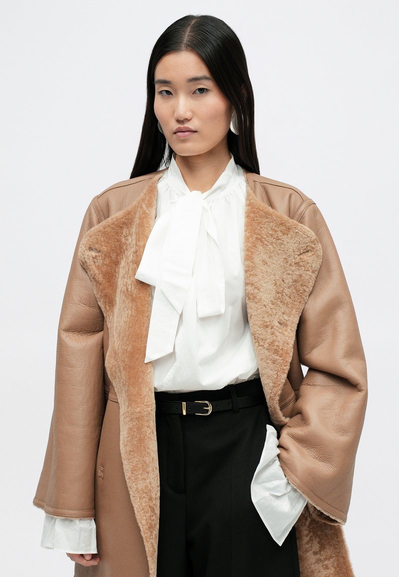 Cappotto in pelle color tan con collo in pelliccia sintetica, camicia bianca con fiocco e pantaloni neri. Presenta maniche larghe e una finitura testurizzata.