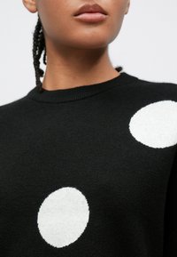 Camisola preta com decote redondo; apresenta grandes bolinhas brancas. Material parece macio com textura suave. Design minimalista.