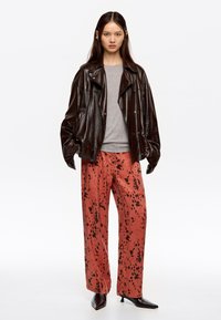Chaqueta de cuero marrón de gran tamaño sobre un suéter gris, combinada con pantalones anchos y estampados en naranja con manchas negras. Tacones negros puntiagudos.