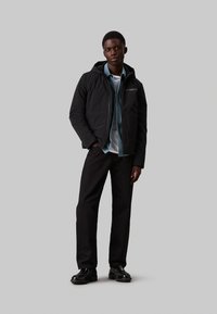 Giacca puffer nera con cappuccio, zip frontale e tasca sul petto sopra una camicia azzurro chiaro. Pantaloni neri a gamba dritta e scarpe nere lucide.