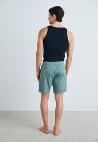Un débardeur noir et un short vert ; le short présente une taille élastique et une texture douce. La silhouette est pieds nus, vue de dos.