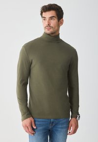 Pulover verde măslin cu guler înalt, dintr-un material moale, cu mâneci lungi și un design ajustat, purtat cu blugi blue. Construcție simplă și fără cusături.