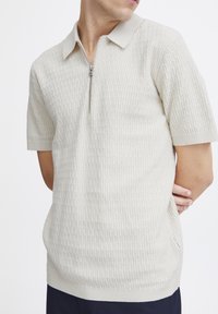 Beige strikket poloskjorte med glidelås på kragen, korte ermer, teksturert overflate og slim fit. Har minimal merkevare på siden.