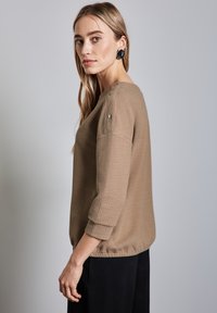 Beige strukturierter Strickpullover mit Dreiviertelärmeln, runder Ausschnitt und dekorativen Knopfdetails an der Schulter.