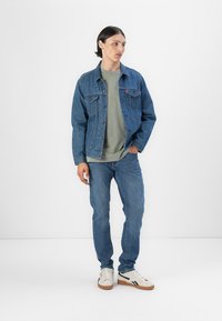Veste en denim bleu moyen, avec fermeture à boutons et poches poitrine, associée à un t-shirt vert et un jean bleu clair. Baskets visibles.