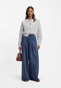 Harmaa, ylisuurikokoinen neuleneuletakki pyöreällä kaula-aukolla ja viidellä napilla; yhdistetty löysiin, korkeavyötäröisiin sinisiin farkkuihin ja pieneen ruskeaan käsilaukkuun.