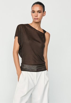 Donna che indossa un top asimmetrico senza maniche marrone scuro e pantaloni bianchi a vita alta e gamba larga, in piedi con le mani nelle tasche.