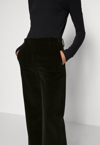 Personne portant un haut noir à manches longues et un pantalon en velours côtelé noir taille haute, les mains dans les poches, devant un fond uni.