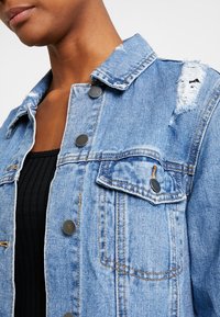 Světle modrá džínová bunda s opotřebovanými detaily, čtyřmi předními knoflíky a dvojitými náprsními kapsami. Texturovaná látka s klasickým límcem.