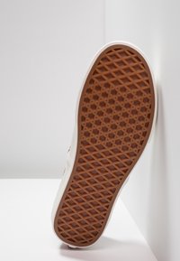 Semelle d'une basket de couleur claire présentant un motif de bande de roulement en forme de gaufre marron, positionnée contre un coin blanc entre un mur et le sol.