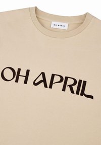 Camiseta beige con escote redondo, que presenta el texto "OH ABRIL" en letras gruesas de color marrón oscuro en el frente. Textura de tela suave.