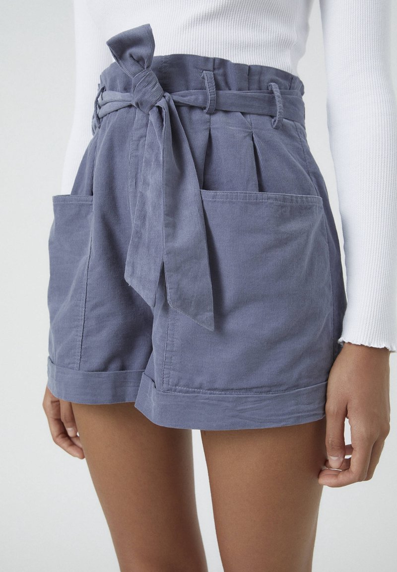 Short en velours côtelé bleu taille haute avec grandes poches avant, ceinture en tissu nouée en nœud, porté avec un haut blanc à manches longues côtelé.