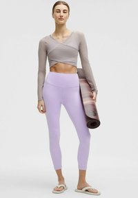 Top a maniche lunghe marrone chiaro, leggings lavanda e sandali bianchi. Tiene un tappetino da yoga arrotolato con un motivo a gradiente in diverse tonalità di marrone e crema.