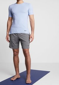 Camiseta atlética de manga corta en azul claro con cuello redondo; combinada con pantalones cortos gris jaspeado, ambos con el logo de Nike. El modelo está de pie sobre una estera azul.