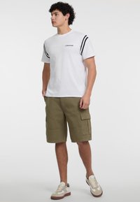 Hvid kortærmet t-shirt med sorte striber på skuldrene, prydet med "Guess Jeans" logo, sat sammen med olivengrønne cargo shorts og sneakers.