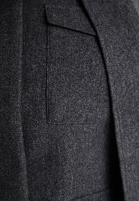 Dunkelgrauer Wollmantel mit strukturierter Oberfläche, der mit einer einzelnen Brusttasche und einem strukturierten Revers ausgestattet ist. Eine Nahaufnahme hebt die Stoffdetails hervor.
