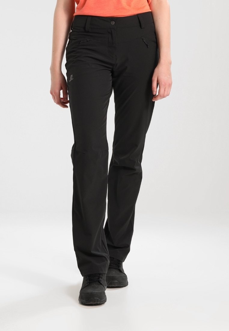 Personne portant un pantalon d'extérieur noir avec des poches zippées et des chaussures de randonnée noires, se tenant devant un fond uni.