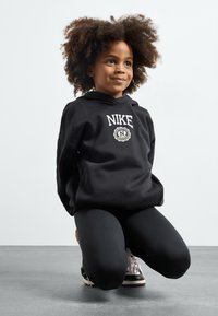 Melnā kapuce ar baltu Nike logotipu, apvienota ar melnām legingu un melnām kurpēm. Modelim ir cirtaini mati un viņš ir ceļos.