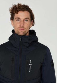 Marinblå pufferjacka med huva, som har dragkedjefickor, texturerade paneler och en logotyp på bröstet. Tillverkad av vattentåligt material.