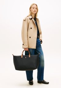 Γυναίκα που φοράει μπεζ παλτό τύπου trench, μπλε τζιν και μαύρα μοκασίνια, κρατώντας μια μεγάλη μαύρη τσάντα tote με καφέ χερούλια.
