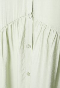Blusa de botão verde-claro, feita de um tecido suave, com detalhes franzidos abaixo dos botões e um brilho sutil, realçando a textura delicada.