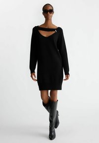 Vestito nero lavorato a maglia con scollatura ampia, dotato di un'apertura e dettagli in metallo silver. Abbinato a stivali neri a girocollo con texture fino al ginocchio.
