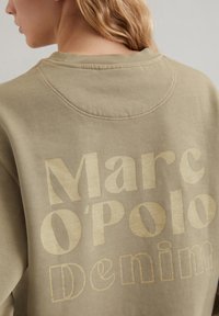 Olivengrüner Pullover mit großen goldenen Buchstaben "Marc O'Polo Denim" auf dem Rücken. Glatte Textur mit rundem Ausschnitt.