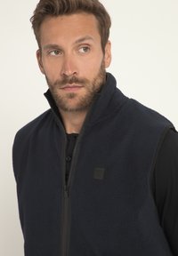 Gilet in pile navy con colletto alto e chiusura a zip. Presenta un tessuto texturizzato e un piccolo logo nero sul petto. Design leggero.