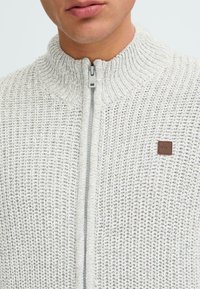 Svetlošedý pletený sveter na zips s vysokým golierom a koženým logom. Obsahuje textúrovaný vzor a kovový zips.