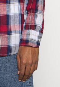 Rood en blauw geruit flanellen shirt met een knoopmanchet. De stof lijkt zacht en textuur te hebben, met een subtiele glans en gelaagde patronen.