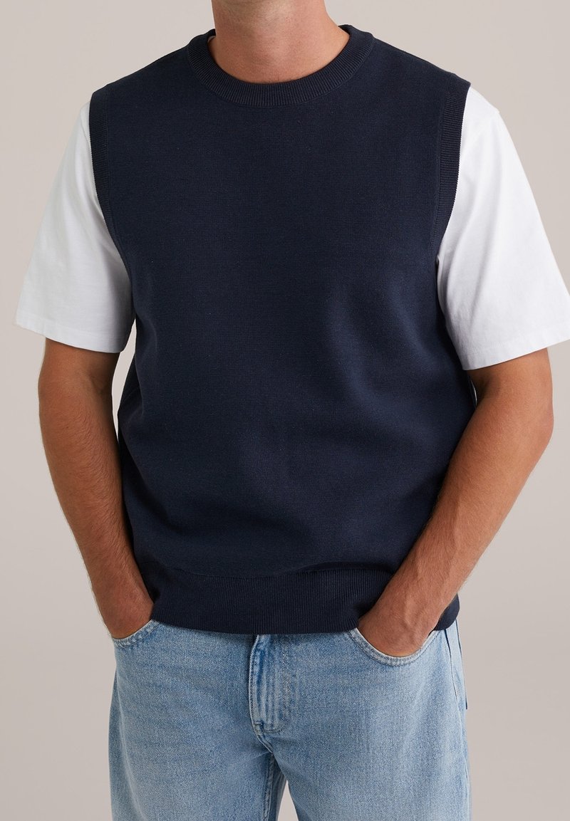 Personne portant un gilet sans manches bleu marine par-dessus un t-shirt blanc à manches courtes avec un jean bleu clair, les mains dans les poches.