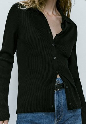 Kobieta w prążkowanej czarnej koszuli na guziki z długim rękawem, częściowo wpuszczonej w niebieskie dżinsy z czarnym paskiem, na białym tle.