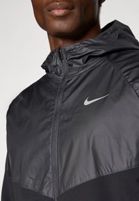 Czarna kurtka Nike z zamkiem błyskawicznym z przodu, z błyszczącą górną częścią i matową dolną częścią. Zawiera kaptur i małe odblaskowe logo.