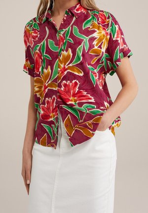 Kobieta w koszuli z krótkim rękawem, zapinanej na guziki, z czerwono-pomarańczowo-zielonym kwiatowym wzorem, lekko wsunięta w białą spódnicę, ręka w kieszeni.