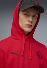 Rode hoodie met trekkoorden, voorzien van een zwart Puma-logo en een AC Milan-embleem. Draagt een zwarte pet. Soepele stoftextuur.