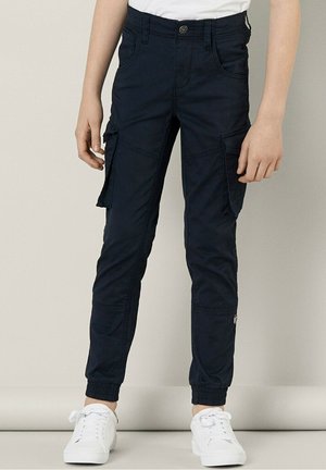 Jeune personne portant un pantalon cargo bleu marine avec des poignets élastiques et des baskets blanches, debout devant un fond clair uni.