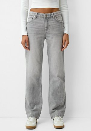 Personne portant un jean taille haute gris clair à jambes droites, un haut blanc à manches longues court et des baskets blanches, sur un fond blanc.